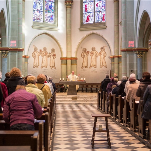 Ein Priester steht an einem Altar in einer Kirche, während Gläubige in den Kirchenbänken den Gottesdienst verfolgen.