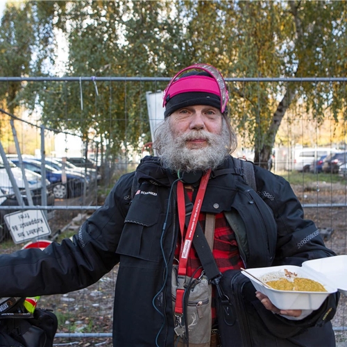 Älterer obdachloser Mann mit Bart und Mütze hält eine Box mit Essen in einem städtischen Umfeld.