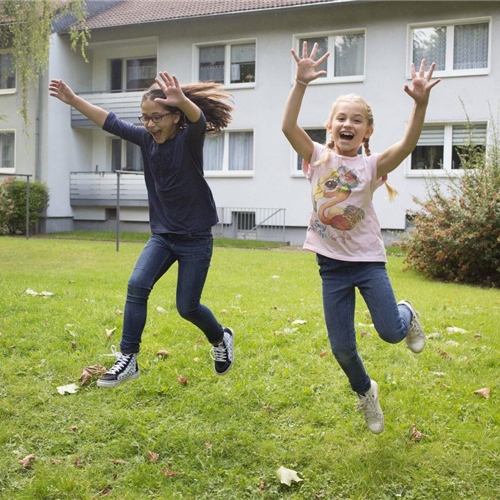 Zwei Mädchen springen fröhlich lachend und die Arme ausgebreitet auf einer Wiese vor einem Wohnhaus.