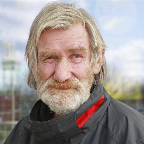 Älterer Mann mit grauem Bart und freundlichem Lächeln steht vor unscharfem Hintergrund.