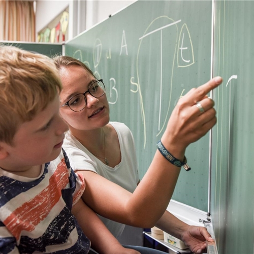 Ältere Schülerin erklärt jüngerem Schüler etwas an einer grünen Tafel im Klassenzimmer.