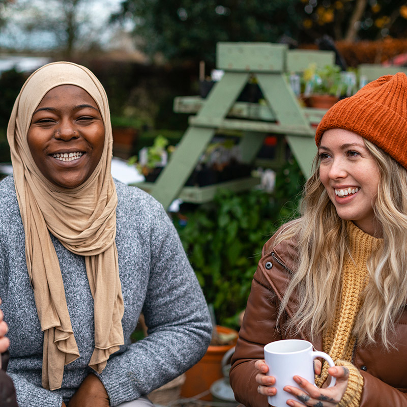 Zwei lächelnde Frauen unterschiedlicher Herkunft sitzen zusammen in einem Garten.