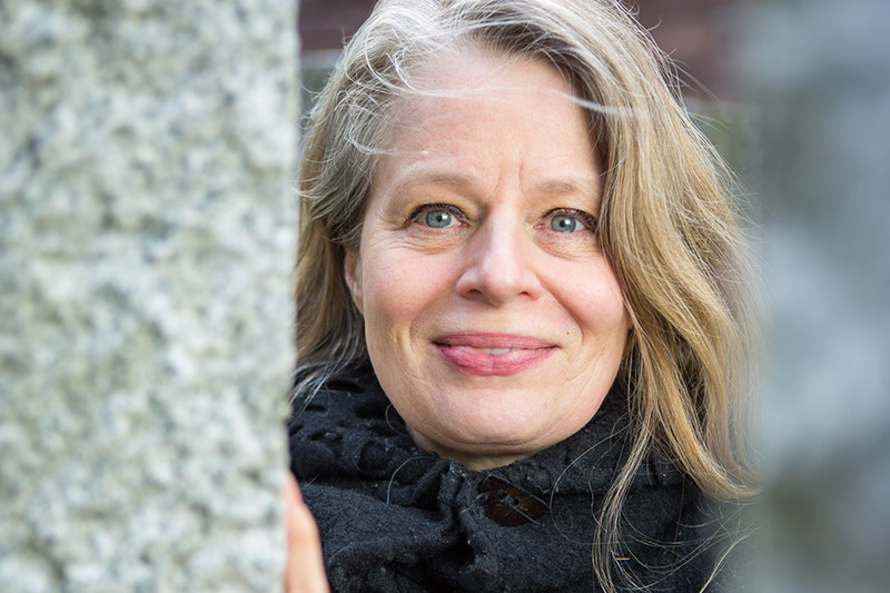 Eine Frau mittleren Alters mit blonden Haaren und blauen Augen schaut hinter einem Baumstamm hervor und lächelt.