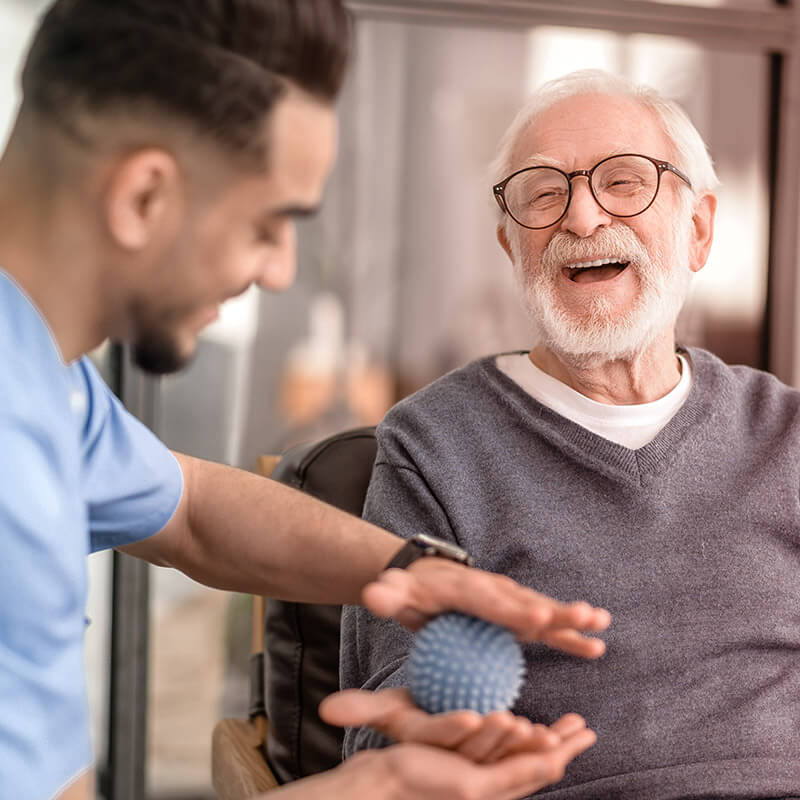 Ein älterer Mann lacht freudig während ein jüngerer Mann ihm seine Hand mit einem Massageball massiert.