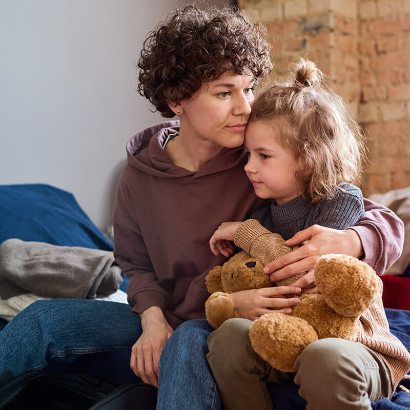 Eine besorgte Frau hält ein kleines Mädchen und ihren Teddybären im Arm.